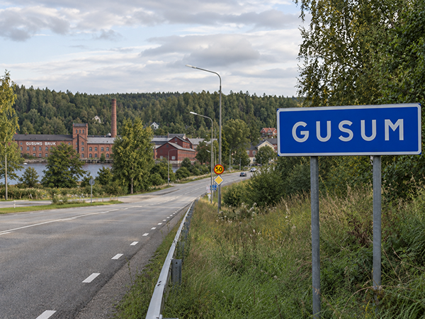 Vägskylt med texten Gusum med det gamla bruksområdet i bakgrunden